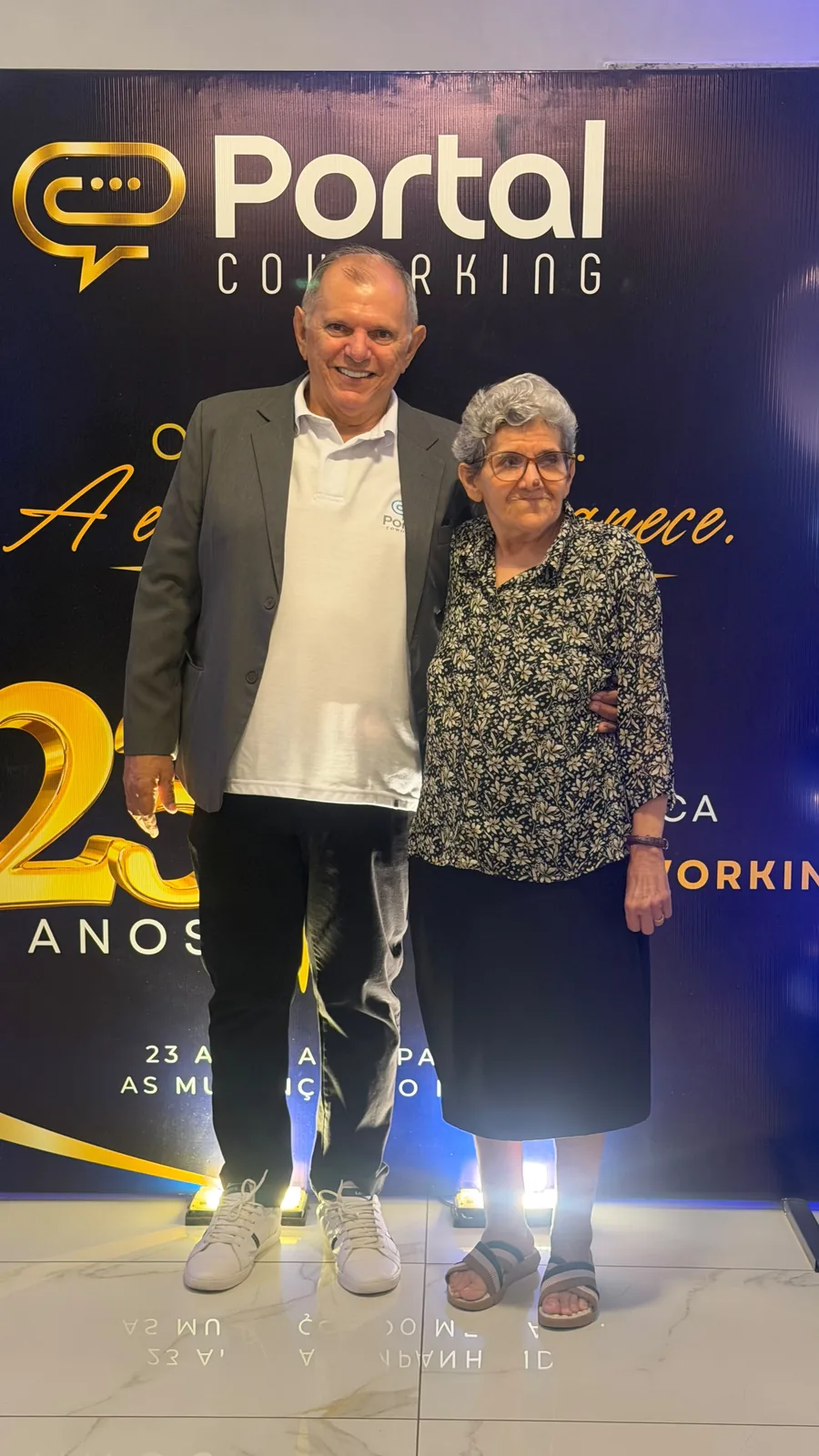 Convidada e anfitrião posam em frente ao painel comemorativo dos 23 anos do Portal